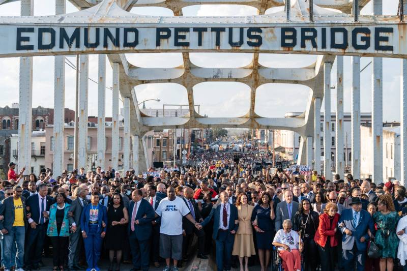 A march of hundreds of people walks across a white bridge painted with the words ‘Edmund Pettus Bridge.’