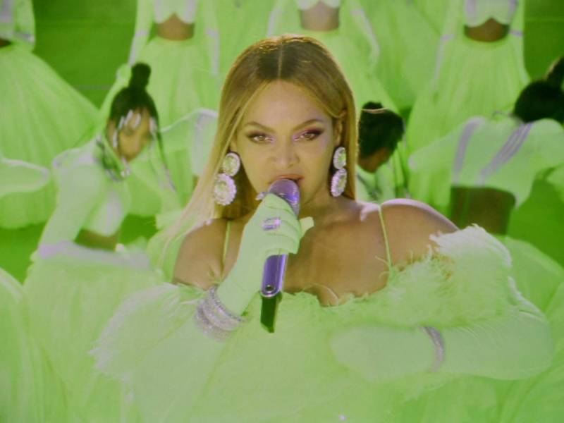 A beautiful Black woman wears an off the shoulder neon green gown and sings into a microphone.