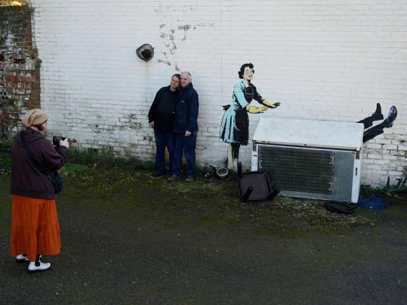 Two people pose for a photo next to a Banksy mural on a white brick wall, depicting a 1950s housewife with a swollen eye, missing a tooth, and shutting a man’s body in a freezer.
