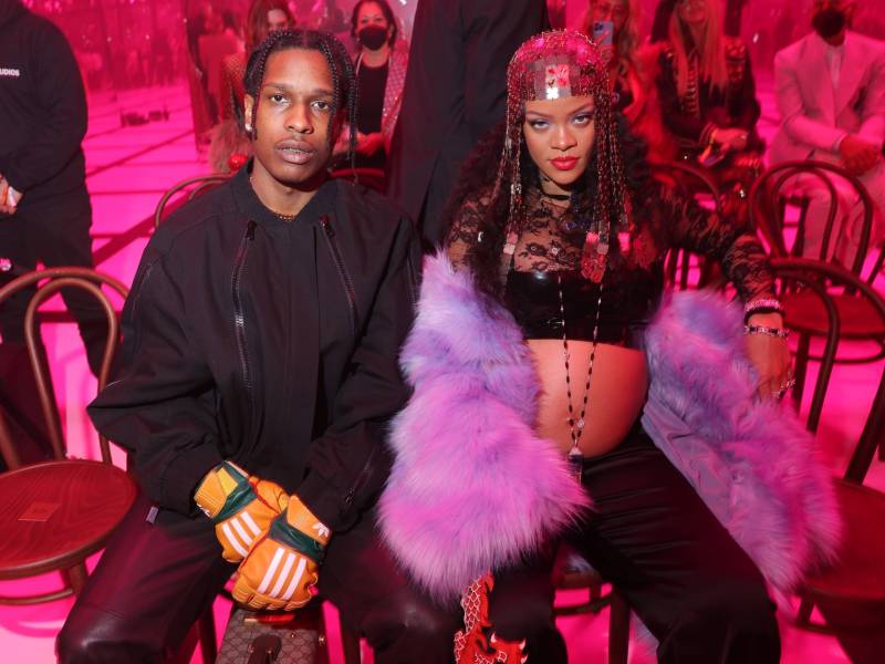 A Black man wearing an all black outfit of jeans, t-shirt and denim jacket sits next to a heavily pregnant Black woman. She is wearing black pants, a black lace fitted crop top and a lilac fake fur stole.