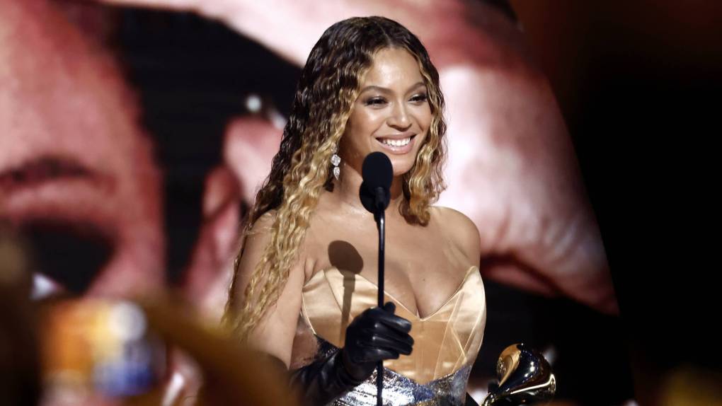A beautiful Black woman with long wavy hair smiles broadly behind a podium microphone, award in hand.