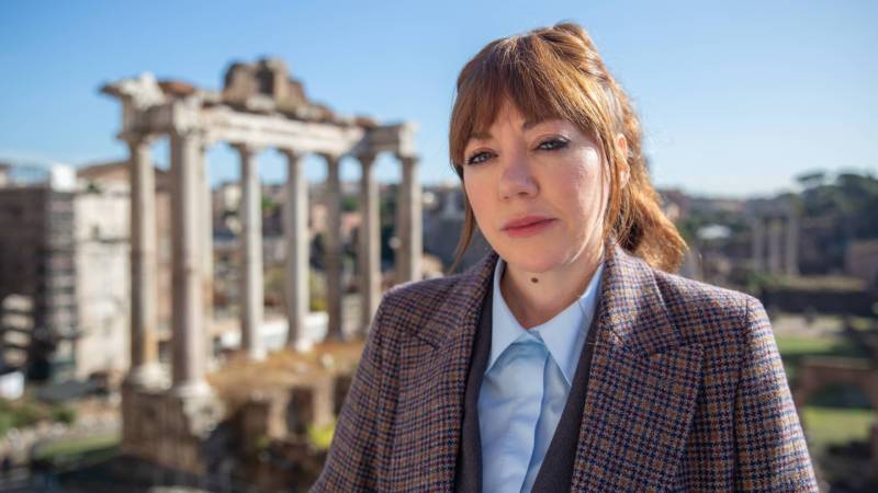 A woman dressed in a smart suit and overcoat, with her red hair pulled back into a ponytail, stands in front of ancient ruins.