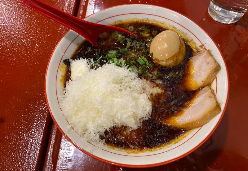A bowl of ramen: red-tinted broth topped with pork chashu, egg, scallions and a flurry of grated Parmesan cheese.