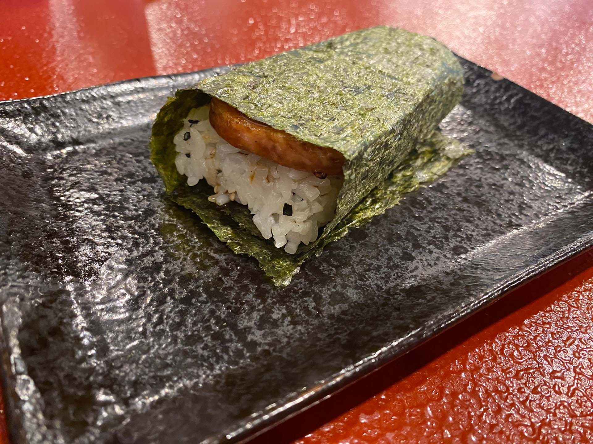 Spam musubi — spam and rice wrapped in a large piece of toasted seaweed — served on a black plate.