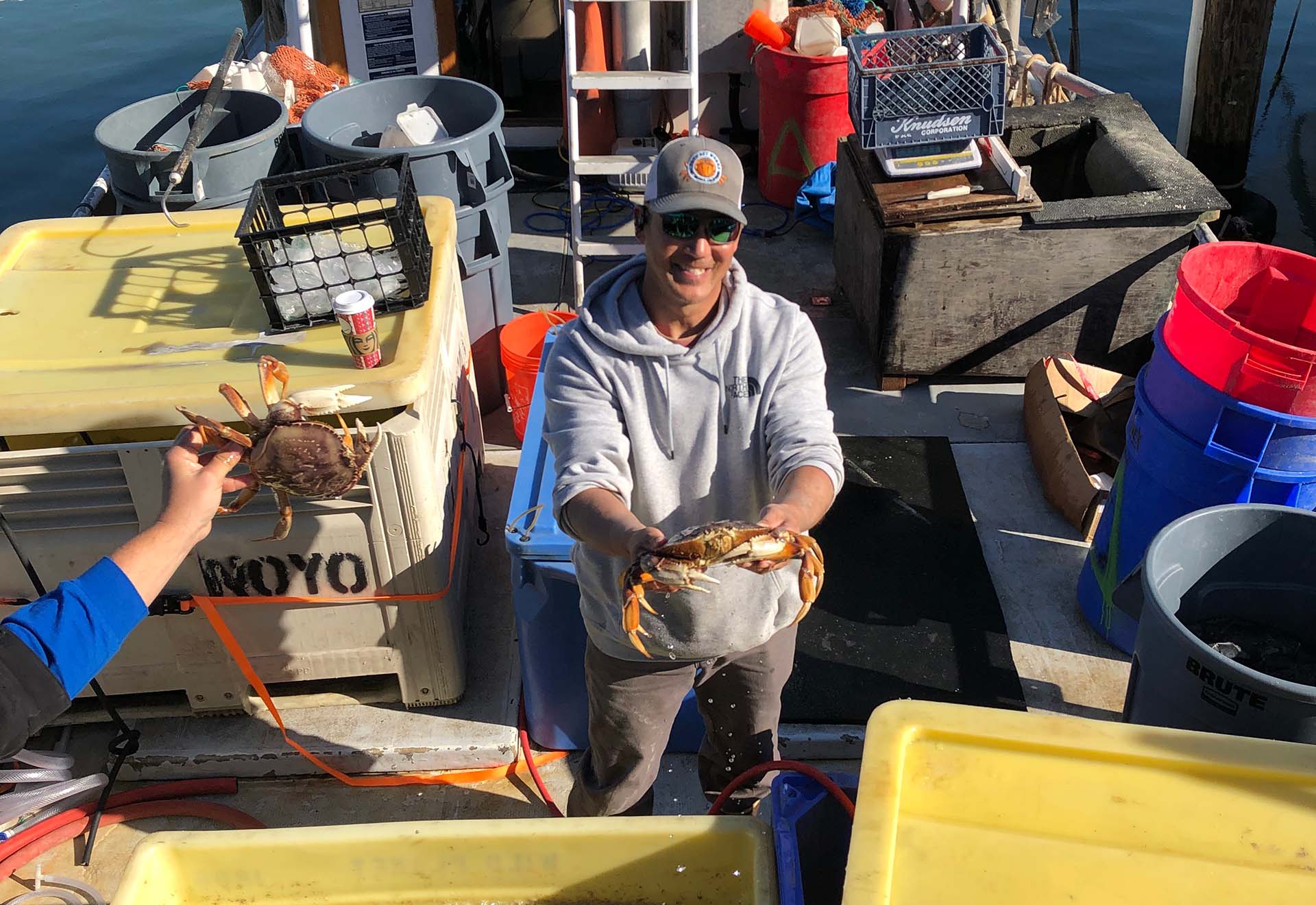 A crab fisherman in a trucker hat smiles as he holds up a live Dungeness crab from the deck of his fishing boat.