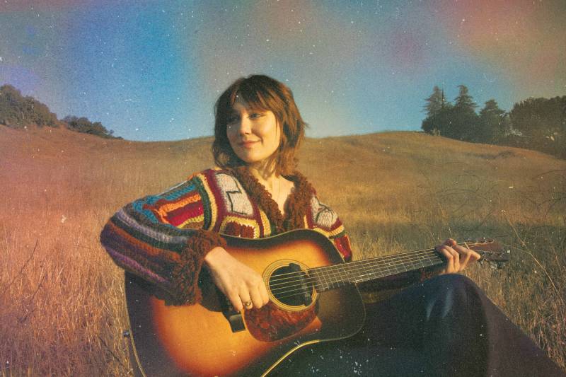 a young white woman in a colorful shirt sits in a field with a guitar, smiling