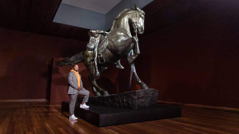 A Black man stands with one foot up on base of a giant brass sculpture of a horse with a man draped over saddle