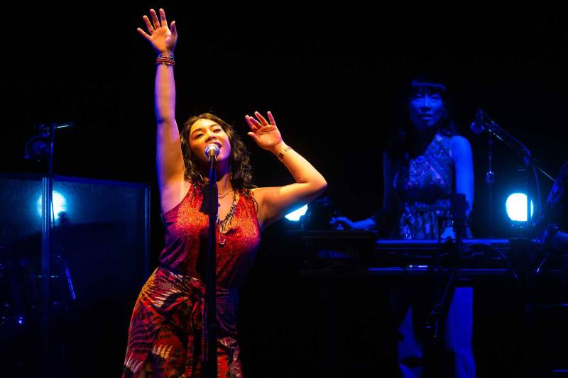 A Cambodian woman in a red dress sings into a microphone while reaching to the ceiling; a keyboardist in dark blue lighting stands behind