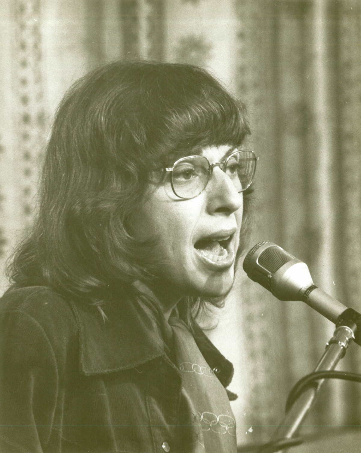 Sepia toned image of white woman in glasses singing into mic