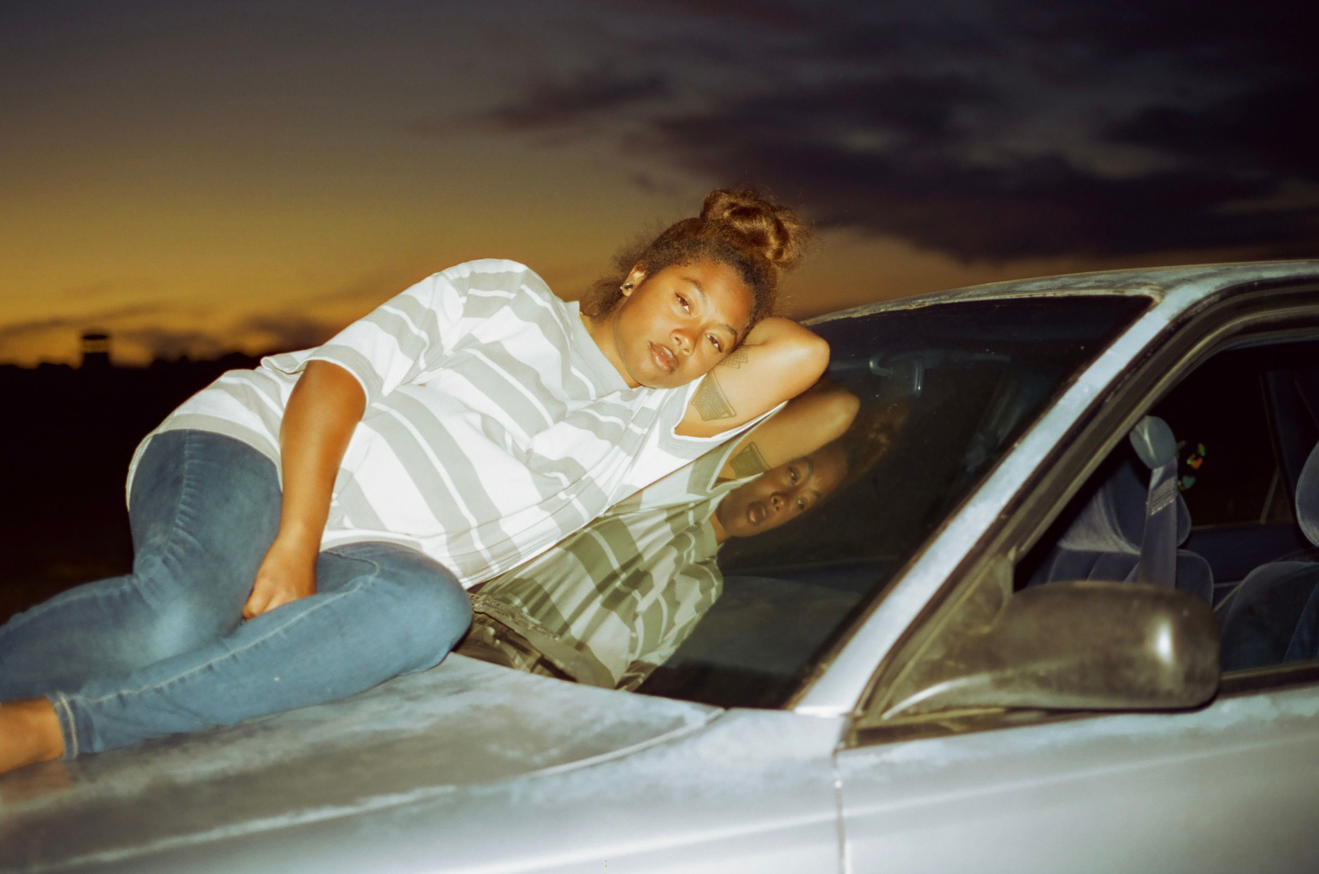 Young Black woman lays on hood of car leaning against windshield