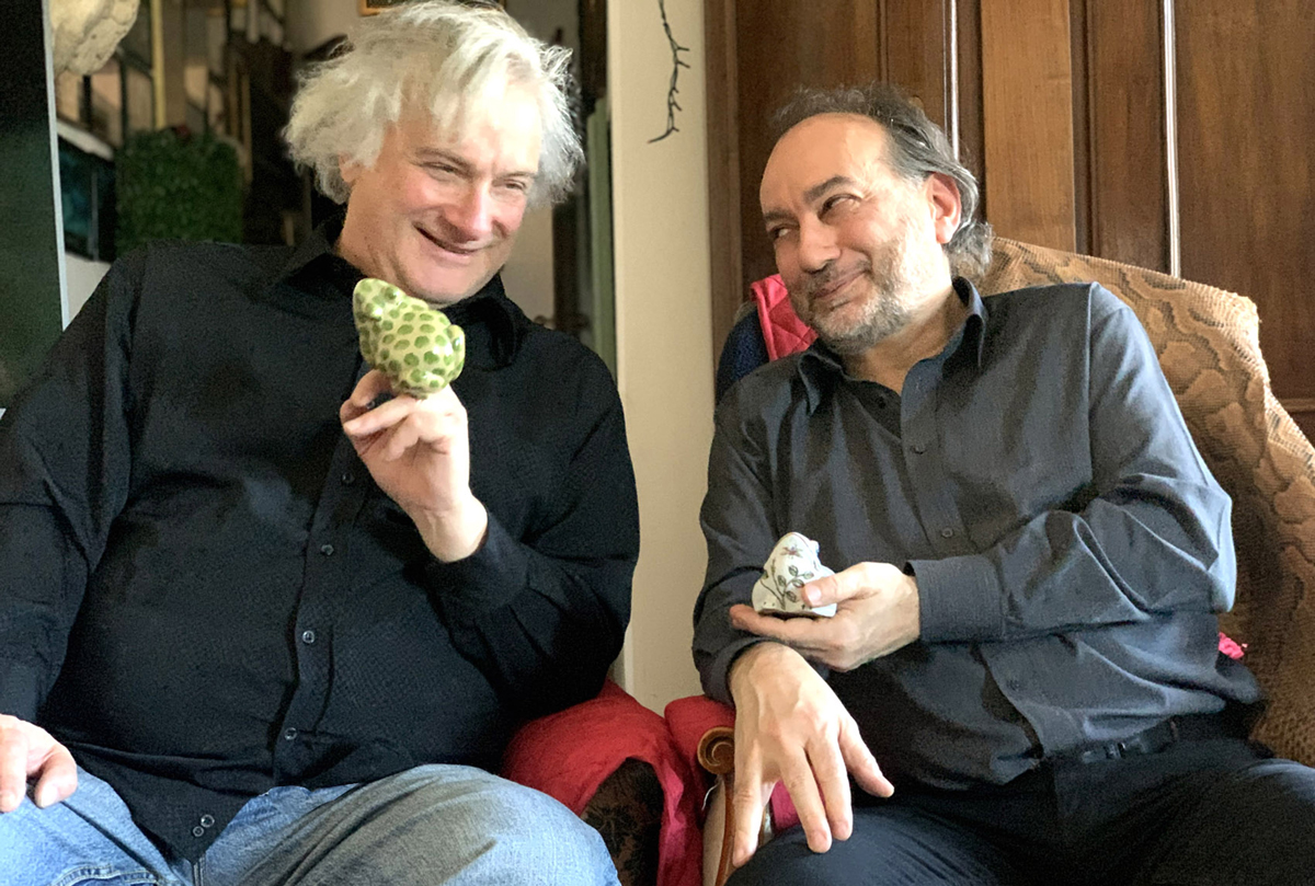 Two white men pose seated, smiling with ceramic frogs in their hands
