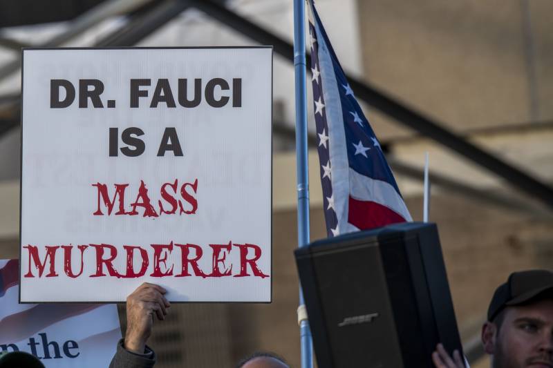 A hand holds aloft a large printed sign that reads "Dr. Fauci is a mass murderer." 