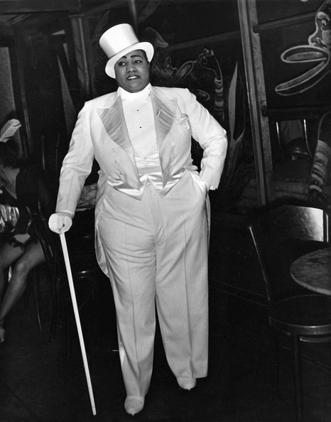 A large Black woman wearing a white tuxedo, top hat and gloves, as she holds a cane.