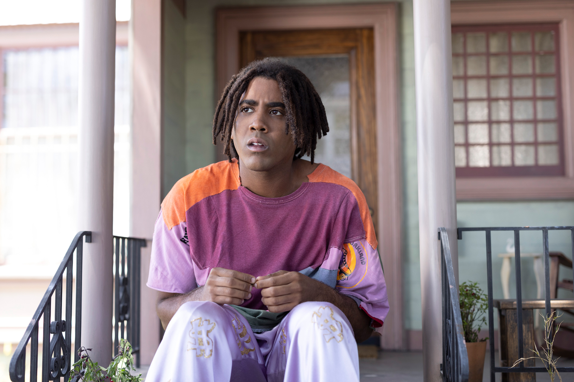 A giant Black man with locs sits on front steps in purple outfit