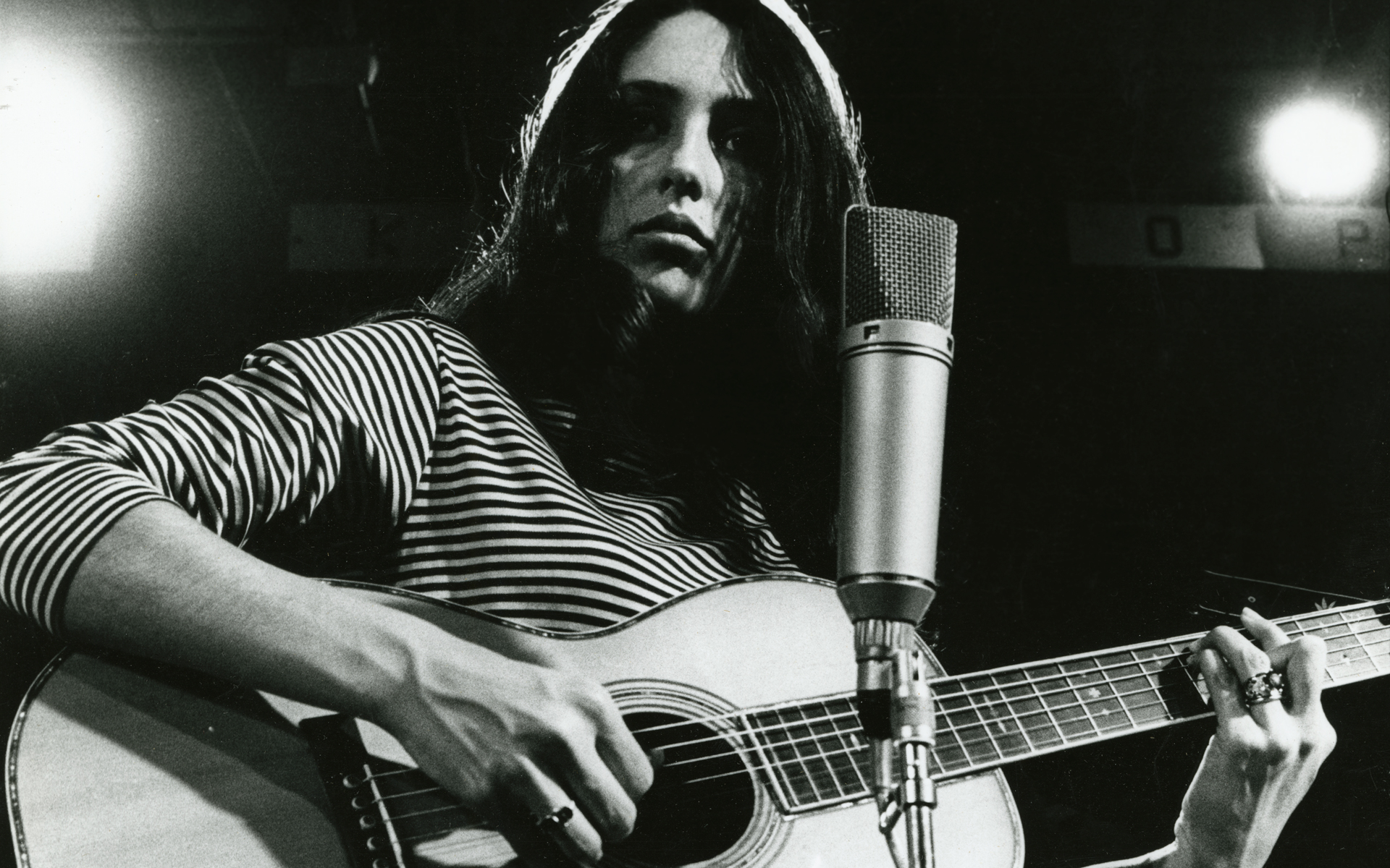 Black-and-white photo of woman in striped shirt playing guitar in front of mic