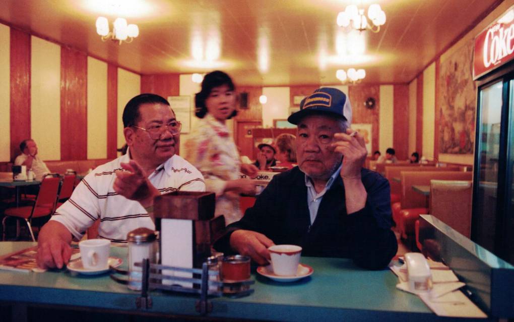 Two older Chinese men sit at a restaurant's diner-style counter while a waitress looks on.