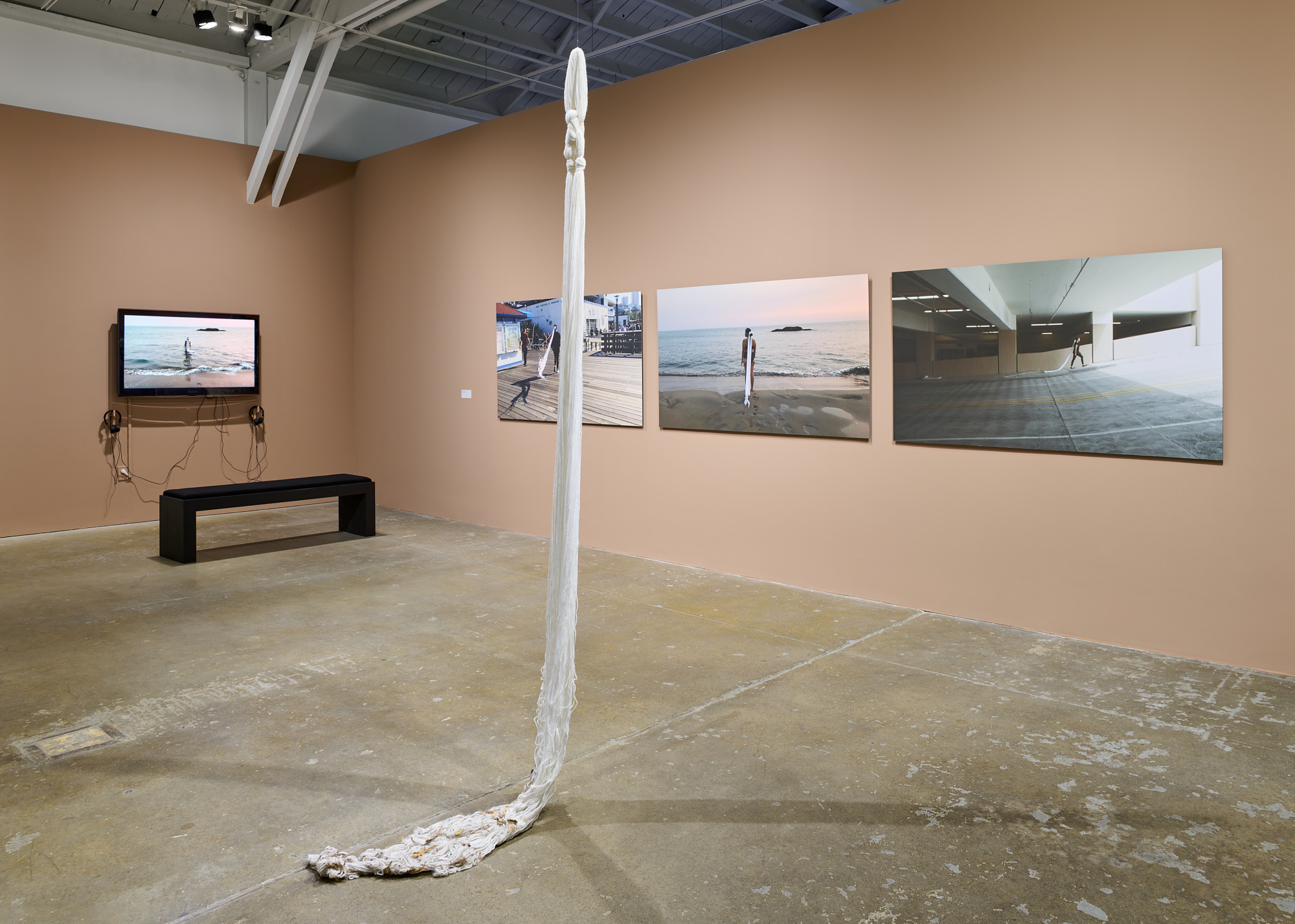 Tan walls with video and three photographic prints, hanging textile sculpture rests on floor