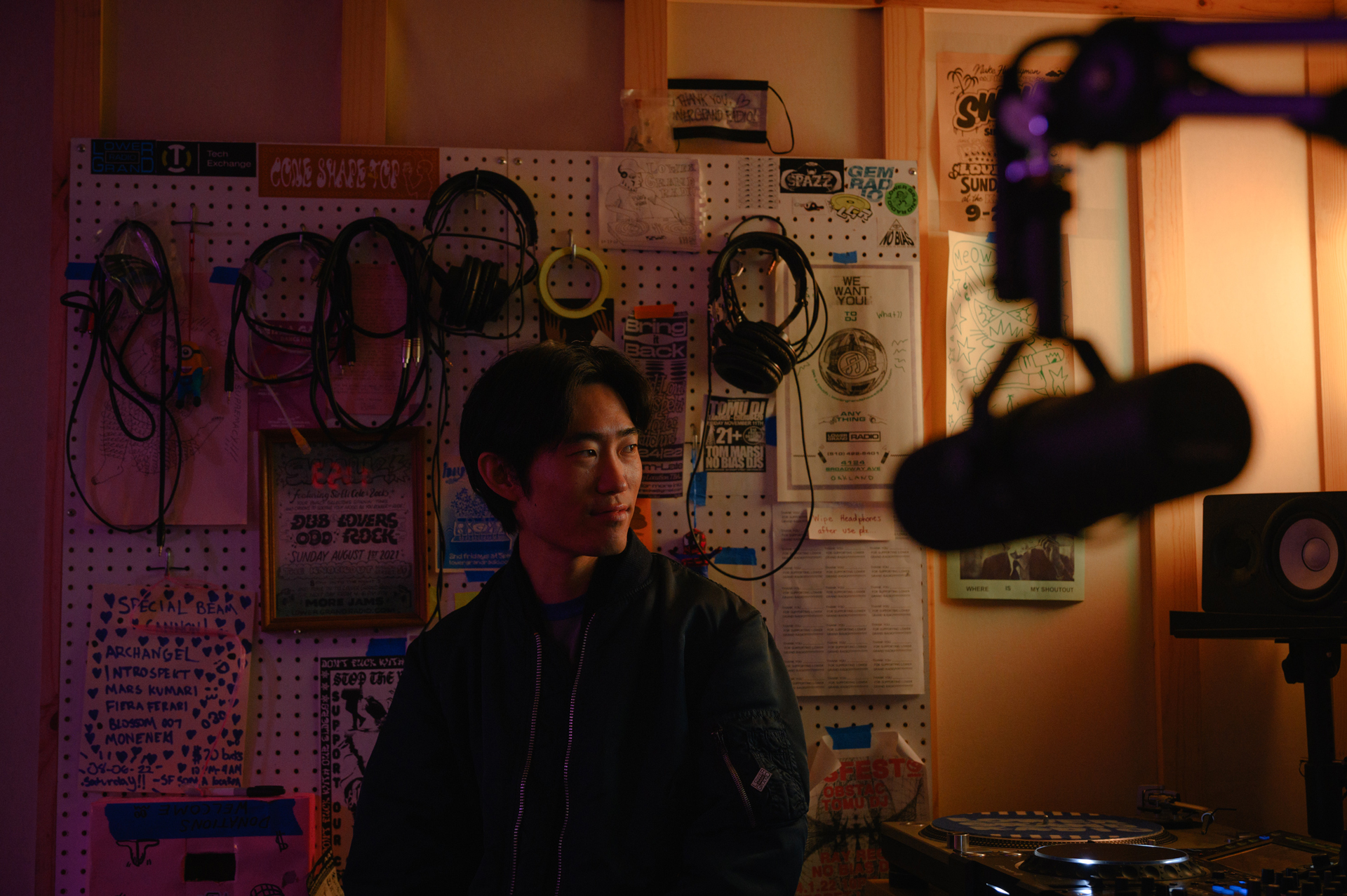 Asian man in hoodie sits in front of pegboard covered with cables and headphones, mic in foreground