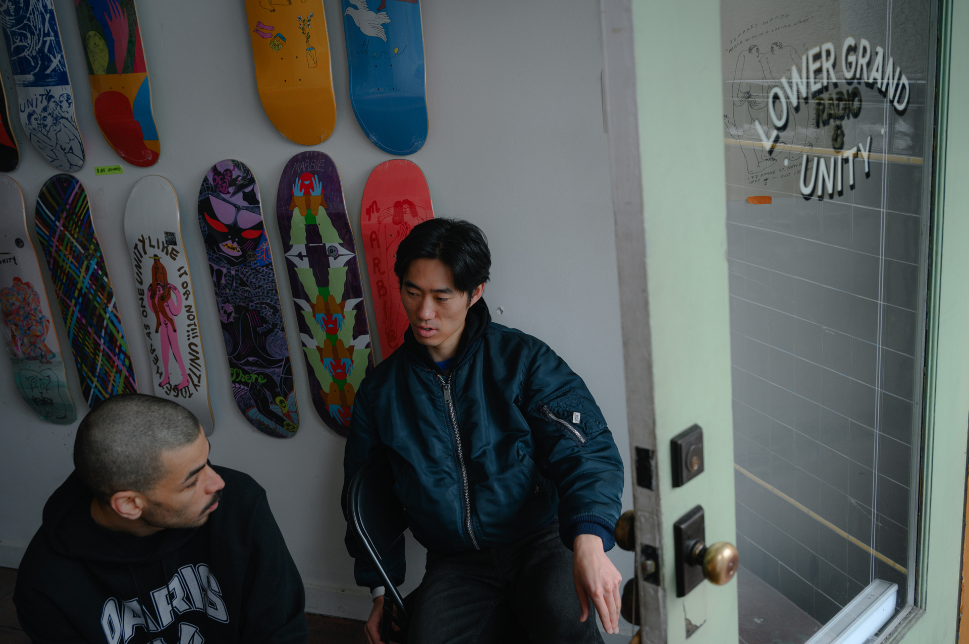 Two men stand in front of wall of skateboards with front door open to right