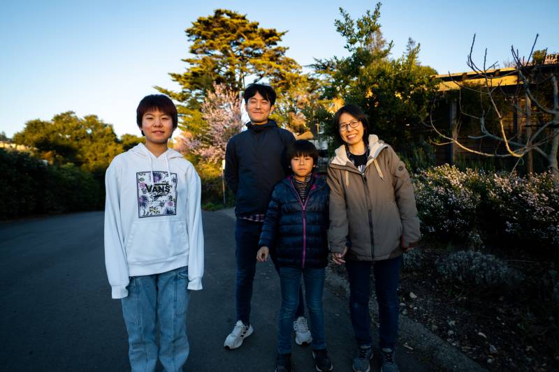 A family of four people, Japanese immigrants, smile for a portrait outside in front of trees: a teenage girl, her father, her younger brother and her mother