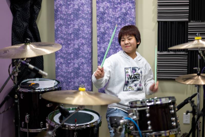 a young Japanese girl with a short haircut in a white sweatshirt smiles while seated at a drum kit in front of a purple acoustic panel in a home studio