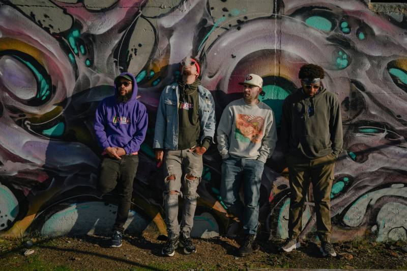 A group of local rappers stands in front of a wall with graffiti in Richmond, CA