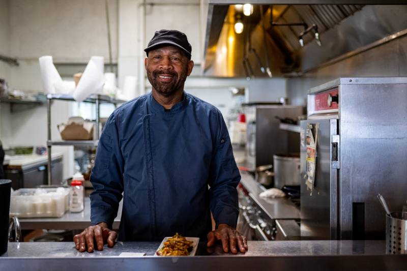 A chef in blue chef's coat smiles inside a restaurant kitchen, a plate of Jamaican food in front of him on the counter.