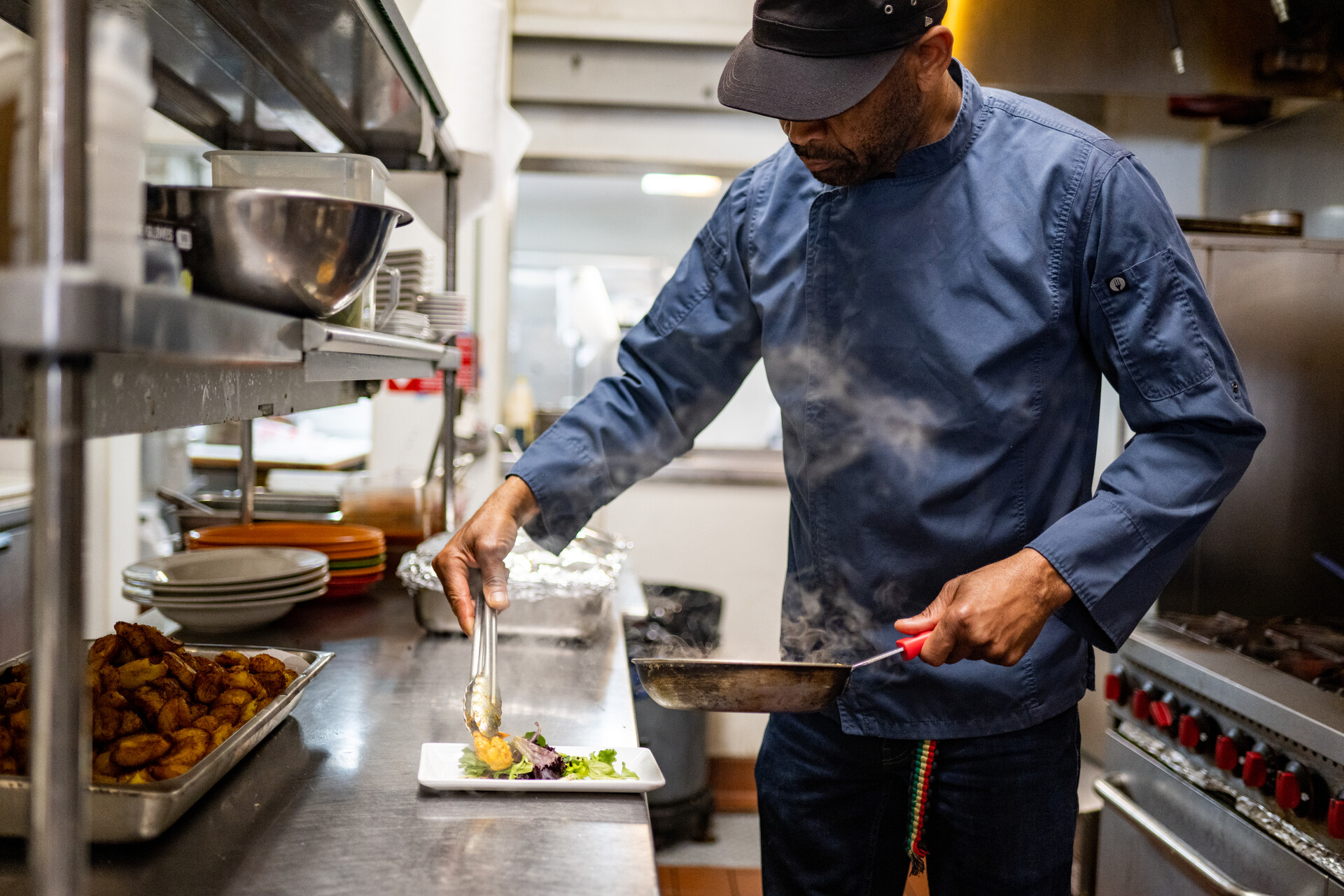 A chef, dressed in a blue chef's coat and black baseball cap, carefully plates an order of curry shrimp.