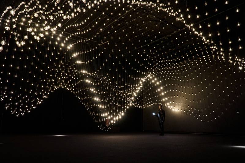 a visitor stands under a massive art installation of tiny lights in a dark room