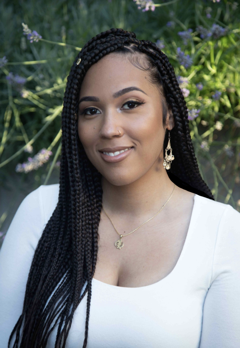 An attractive Black woman with long braids stands in a garden, smiling for the camera. She is wearing a long-sleeved white top and delicate gold earrings and necklace.