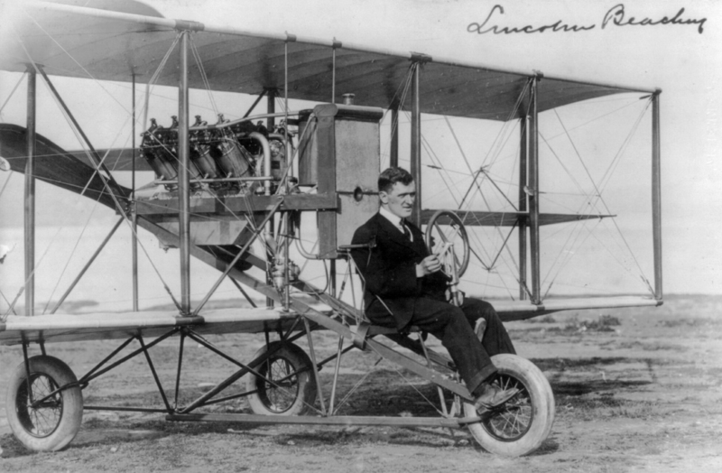 A man wearing a suit sits in a very basic airplane from the early twentieth century.