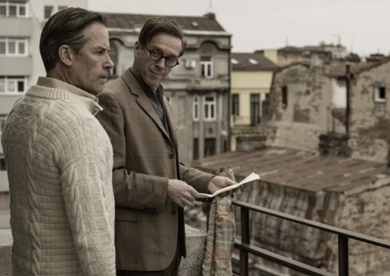 Two men stand on a balcony overlooking European rooftops. One wears a white woolen sweater and a look of concerned. The other is in a brown tweed suit and round spectacles, looking curious.