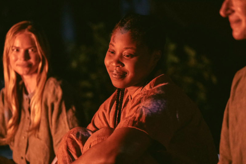 A young Black woman sits in front of a campfire, a serene expression on her face. A white woman sits nearby, a half-smile on her face.