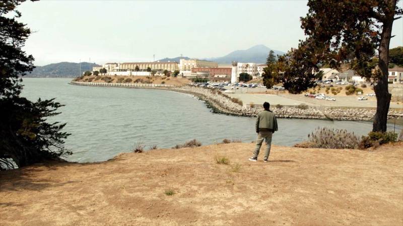 A man gazes toward a large concrete complex that sits near a bay of water.