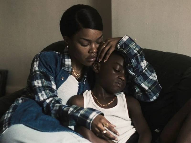A young Black woman wearing a flannel shirt embraces a young, tired-looking Black boy. They are sitting on a couch together in a sparse room.