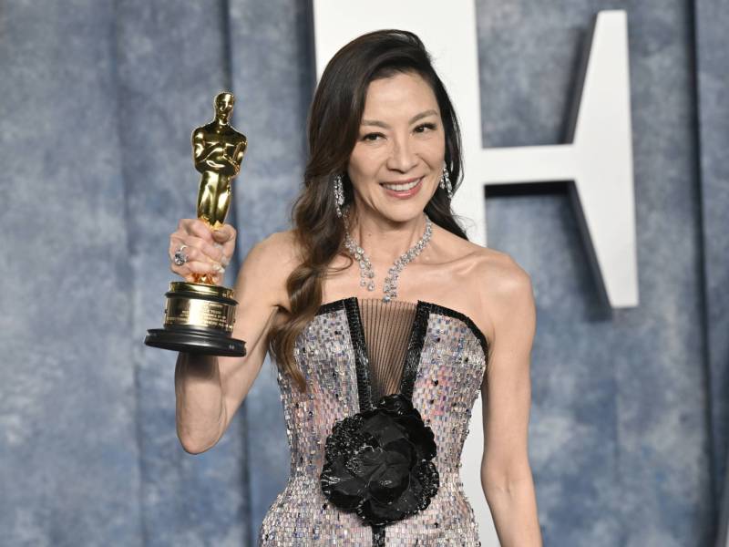 A slender Asian woman wearing a strapless sequined evening gown, smiles and holds an Oscar statuette aloft.