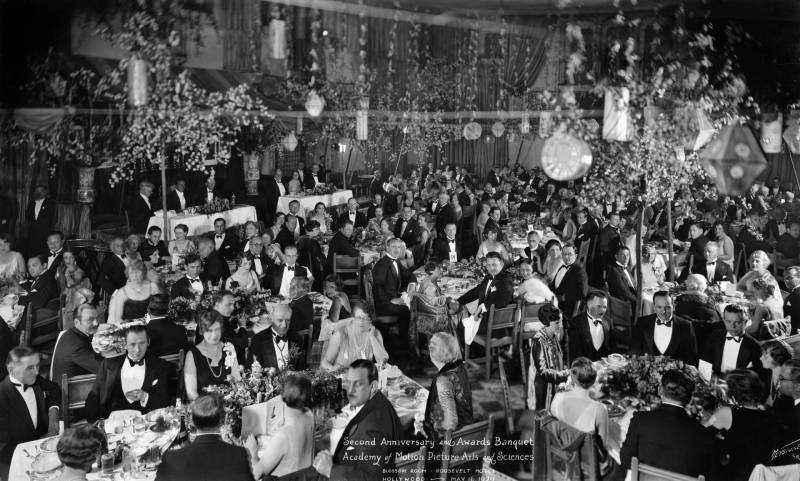 A room of men and women dressed in 1930s evening attire sit at dining tables in a large hall. Festive decorations hang from the ceiling.