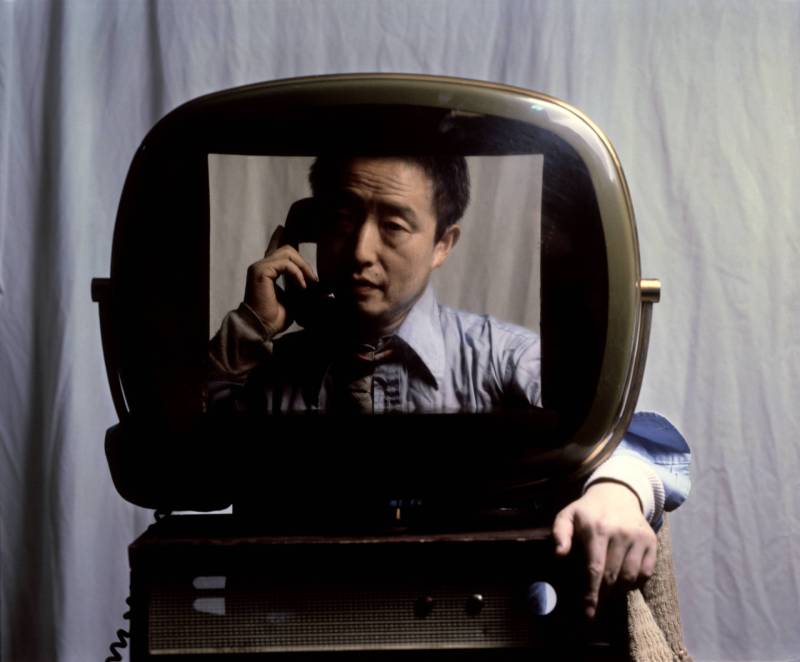 An Asian man sits in front of a white curtain, holding a telephone receiver to his face. He is sitting behind a TV screen, his arm emerging from the side of it.