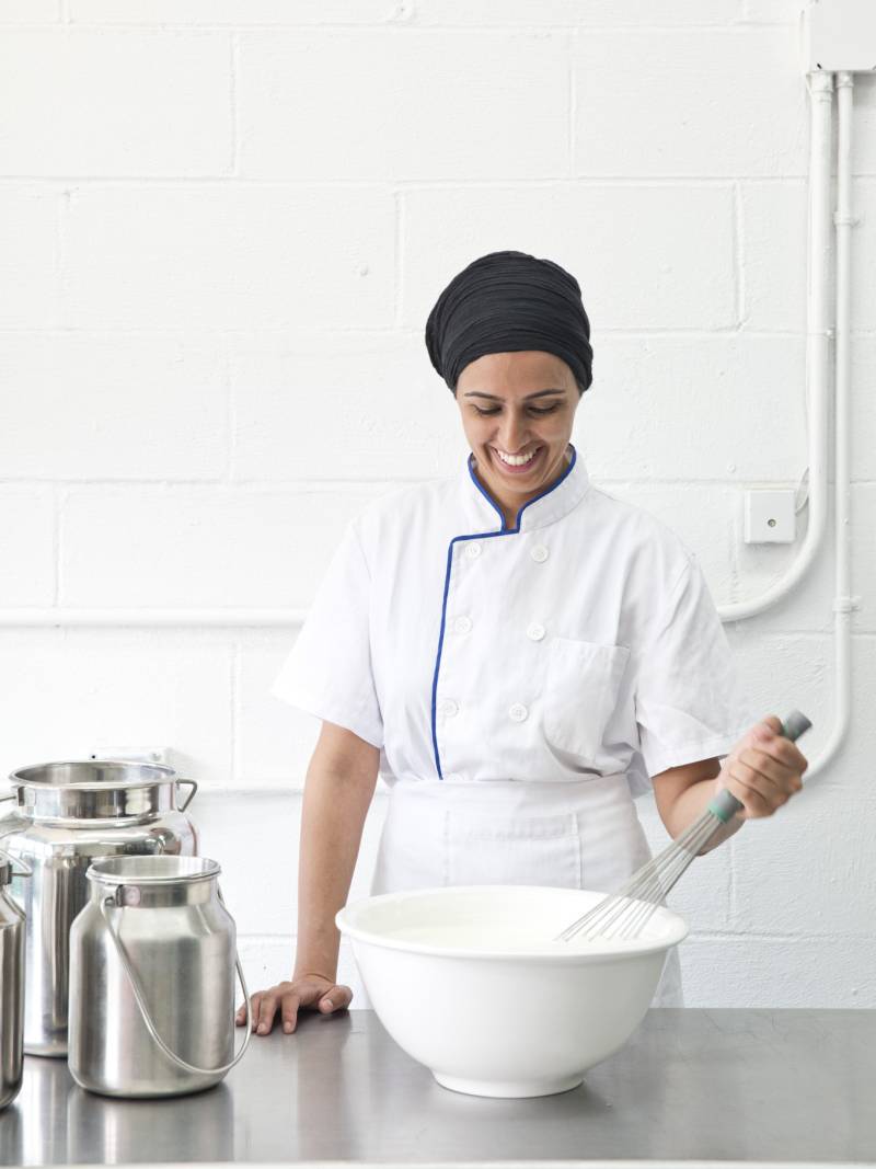 A woman stands in a bright, white kitchen, dressed in crisp white chef's uniform. She is standing over a white mixing bowl smiling.