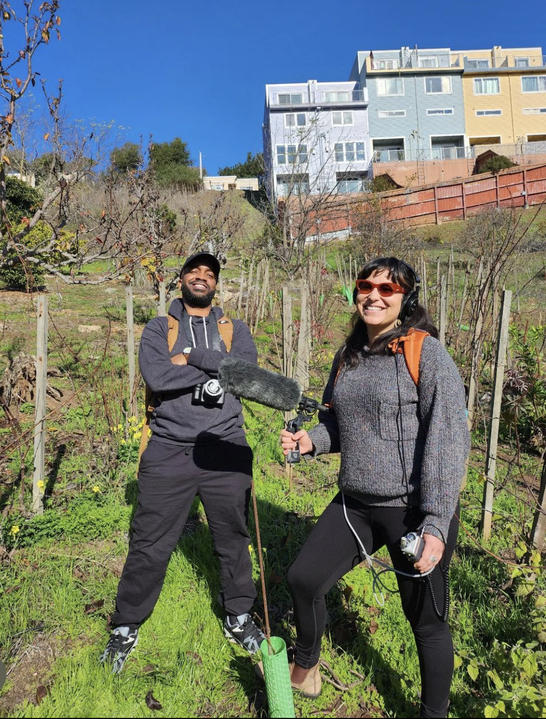 Marisol Medina-Cadena holds a boom mic while Pendarvis Harshaw poses with his camera around his neck, as they stand on a hillside in San Francisco-- the site of the 280 Project's wine vineyard.