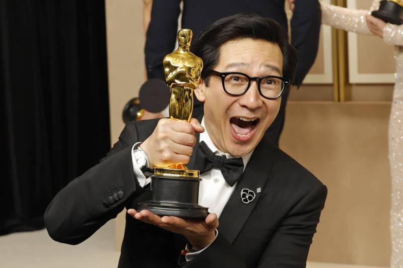 An Asian man wearing black spectacles and a tuxedo holds up an Oscar statuette, jubilant expression on his face.