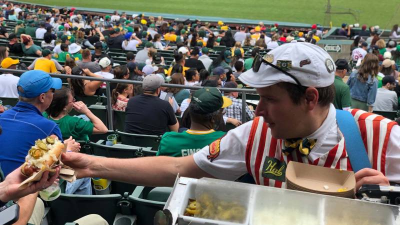 A vendor in a red-striped concessions uniform hands a hot dog to a customer at the Oakland Coliseum.