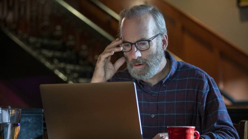 A white man with grey hair and beard stares at a laptop screen looking stressed. One hand is raised and touching the rim of his spectacles.