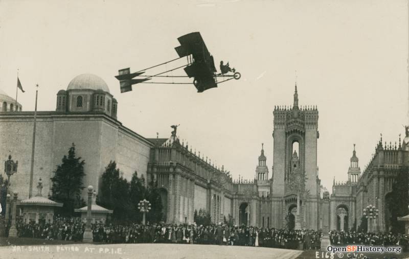 A small wooden airplane resembling a bicycle with wings flies over the heads of gathered crowds. In the background, a palatial building looms.