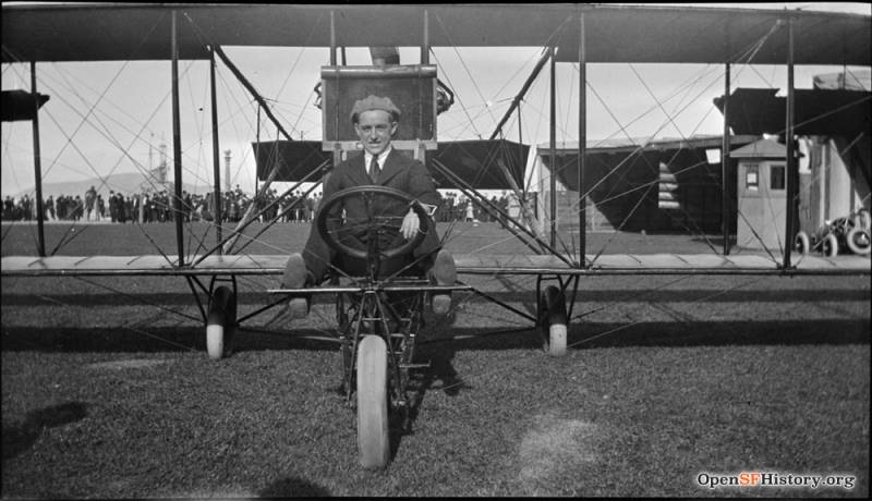 A man in a suit and flat cap pulled backwards sits at the wheel of an early model biplane.