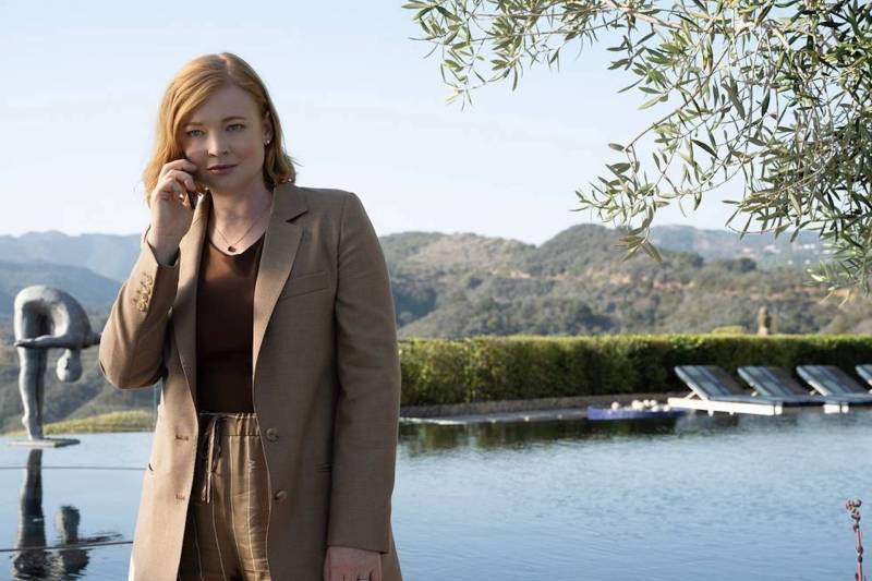A red-haired woman with pale skin stands in front of a picturesque pool and gardens. She is wearing a tan suit and holding a cell phone to her ear.