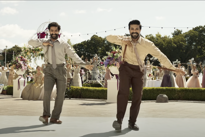 Two Indian men wearing smart trousers, shirts, ties and suspenders perform a dance routine at an outdoor wedding.