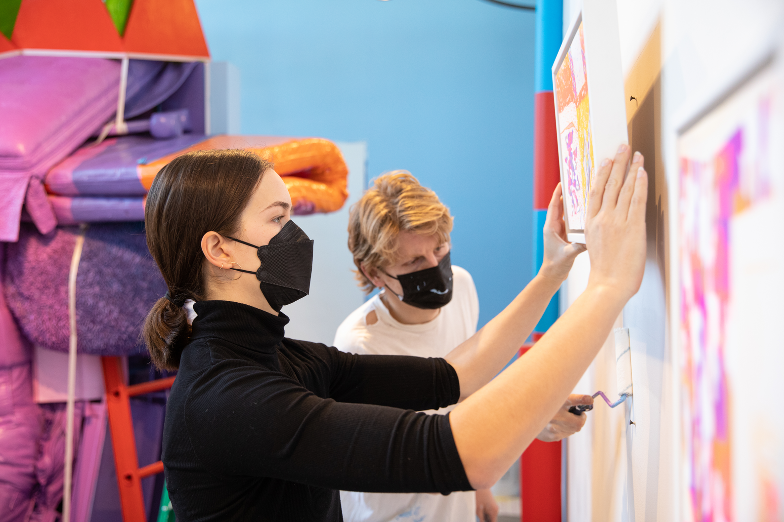 Two students wearing masks install artwork against colorful background
