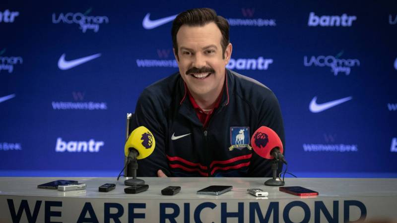 A white man with a mustache sits at a desk facing microphones and smiling broadly. Sports branding is all around him.