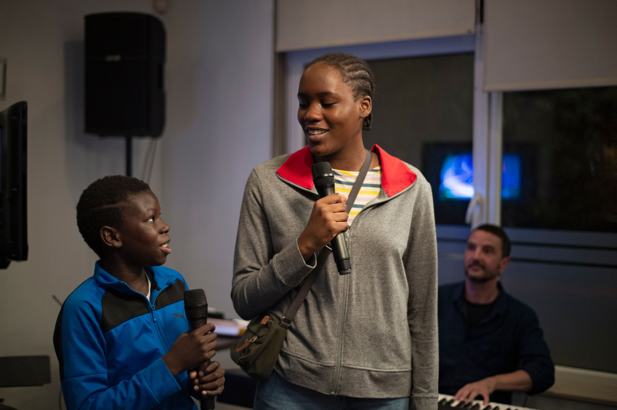 A young Black boy and an older Black girl hold mics and smile at each other with keyboardist behind.
